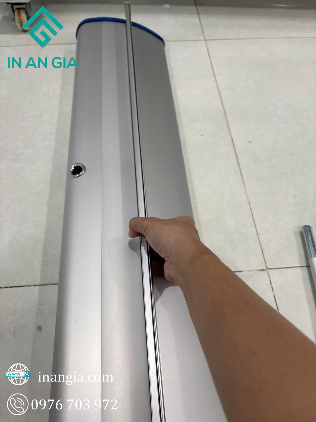 Tay người lắp đặt chân đế standee cuốn nhôm màu bạc của In An Gia trên nền gạch trắng