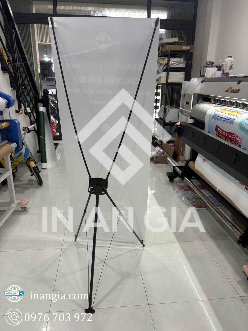 Giá treo standee chữ X bằng kim loại đặt trong xưởng in, phía sau là máy in khổ lớn