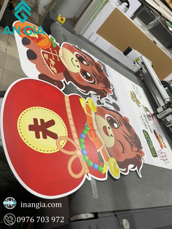 Standee in ấn hình linh vật trâu cầm túi tiền đỏ trang trí Tết trên bàn máy cắt tại xưởng in Angia